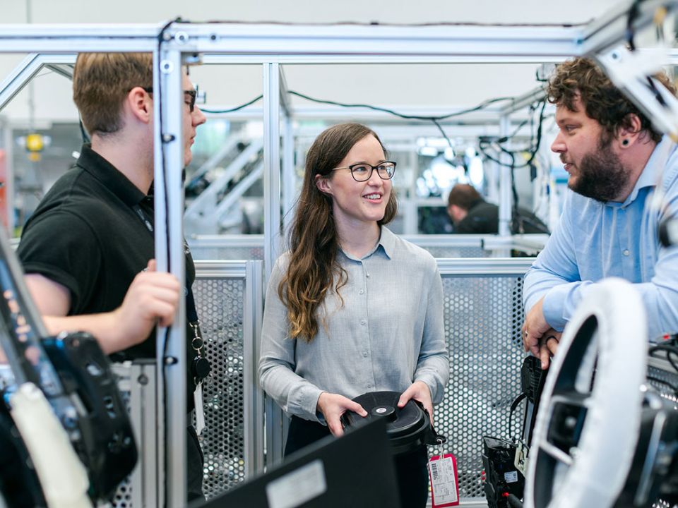 Eine junge Ingenieurin spricht mit zwei Technikern vor dem Fahrgestell eines Autos in einer Produktionshalle