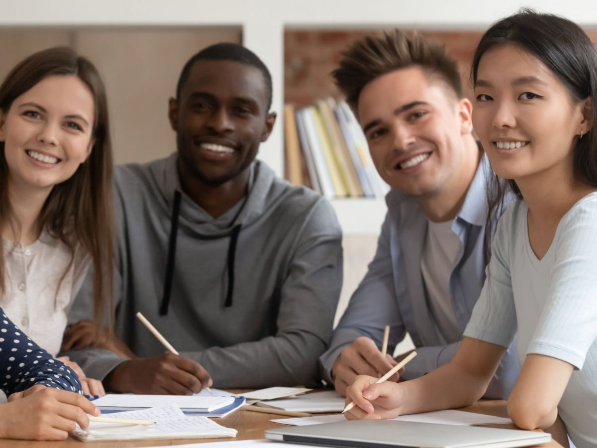 Eine Gruppe von multiethnischen Personen bearbeitet gemeinsam an einem Tisch Unterrichtsmaterial und schaut in die Kamera
