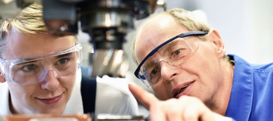 Ein Auszubildender und sein Meister schauen auf eine Maschine, die der Meister dem Auszubildenden erklärt
