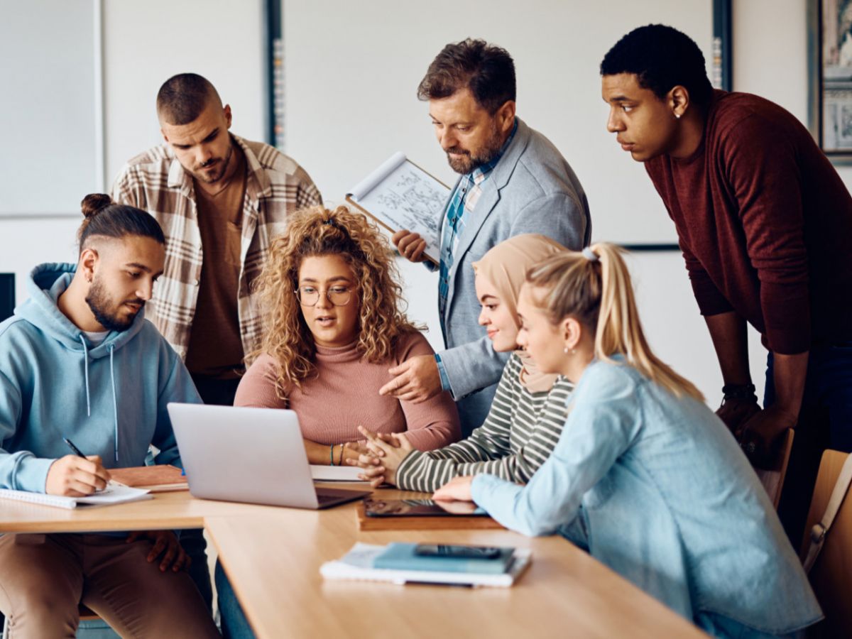 Eine Gruppe von Lernenden sitzt im Unterrichtsraum vor einem Laptop, der Lehrer steht bei ihnen und erklärt