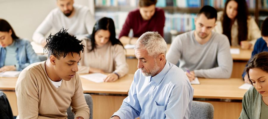 In einem Klassenraum sitzen erwachsene Lernende hintereinander in Tischreihen, ein Lehrer sitzt neben einem Schüler und erklärt ihm ein Thema